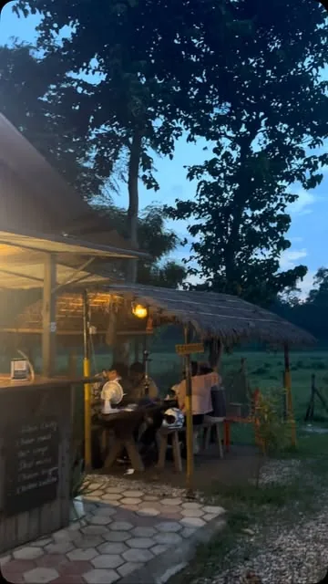 A table under the thatch at dusk, lit by a hanging lantern, chalkboard on the wall.